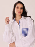 A woman wearing a white long-sleeve blouse with blue and white striped accents on the inner collar placket and the chest pocket, posed against a plain light beige studio background.
