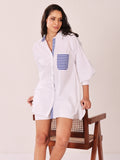 A woman models a white oversized button-up shirt with blue and white striped accents on the collar, placket, and chest pocket, standing next to a wooden chair against a plain light background.